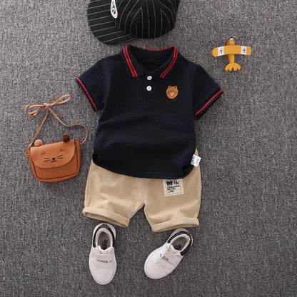Infant outfit with navy polo shirt, beige shorts, white shoes, striped cap, and bear purse