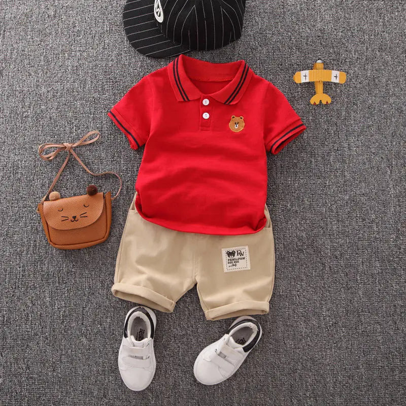 Infant summer outfit with red polo shirt, beige shorts, white shoes, bag, and cap on grey background