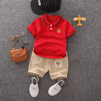Infant summer outfit with red polo shirt, beige shorts, white shoes, bag, and cap on grey background